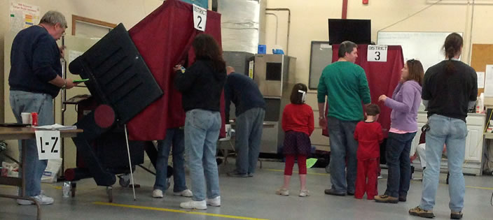 Two poll workers and a voter inside the voting booth. A line of voters waits to check in.